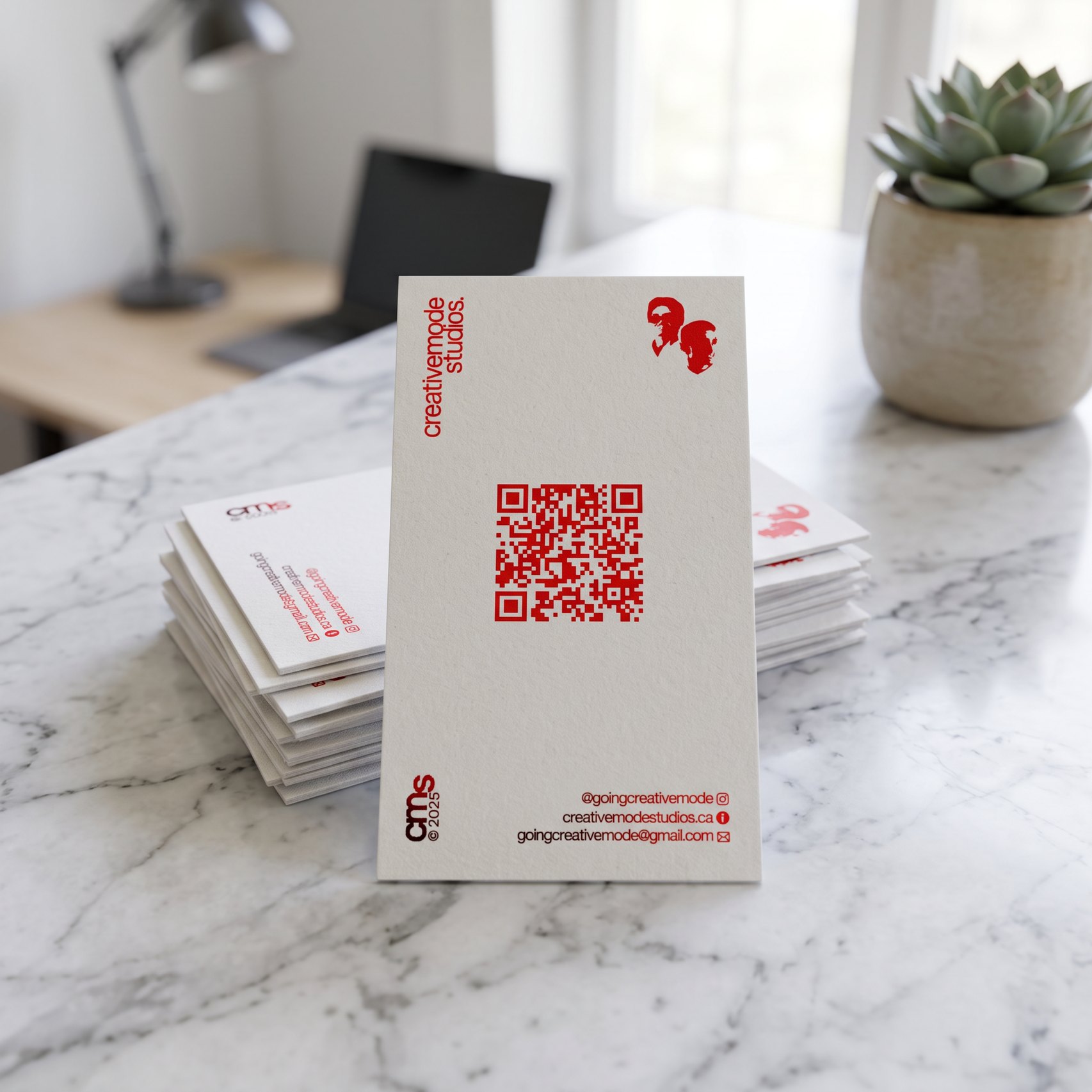 CreativeMode Studios business card standing on a marble desk, with a red QR code and the founders' mark